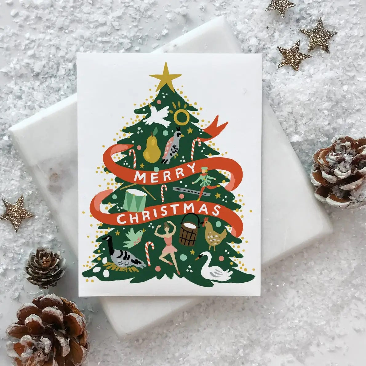 Christmas card with a decorated tree and 'Merry Christmas' text, surrounded by decorative items on a textured surface | Idlewild Holiday Card | 12 Days Tree, Made in the USA