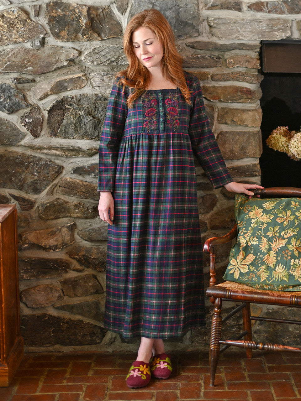 Woman in a plaid dress standing in front of a stone wall with a fireplace.