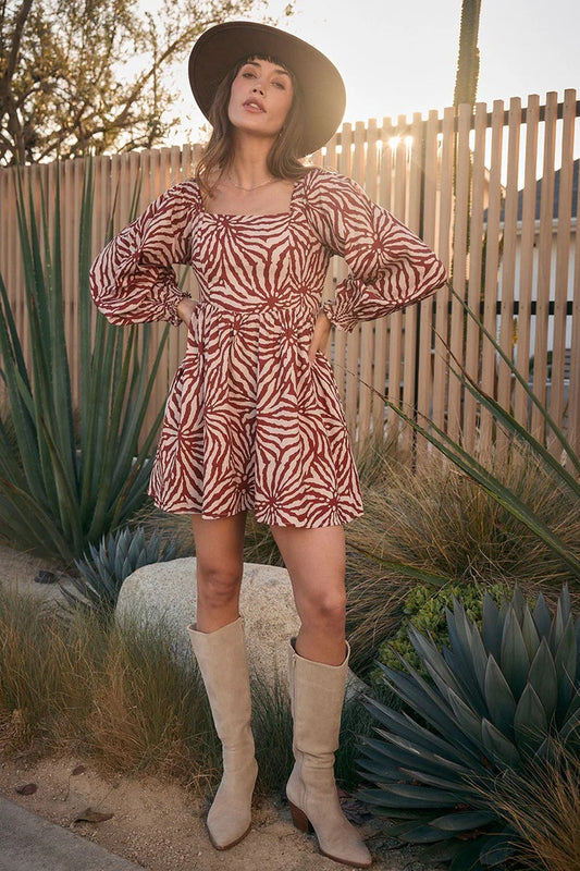 Woman in a zebra print dress and beige boots standing in a desert-like setting with cacti and a wooden fence.