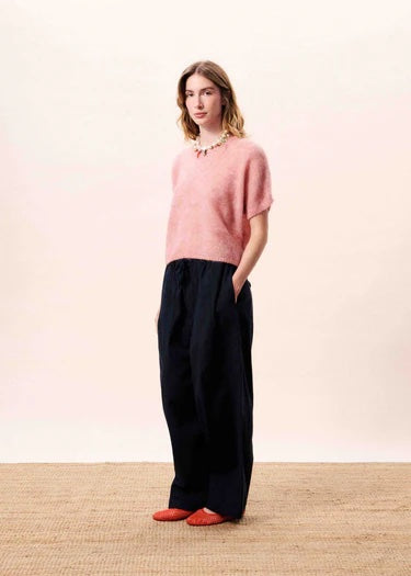 Woman wearing a pink sweater and black pants standing against a plain background