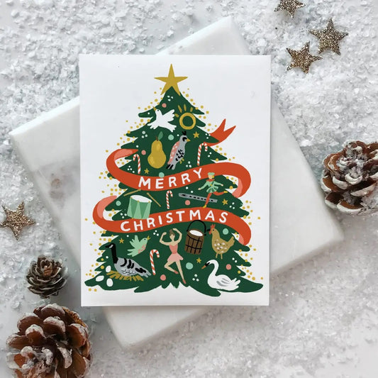 Christmas card with a decorated tree and 'Merry Christmas' text, surrounded by decorative items on a textured surface | Idlewild Holiday Card | 12 Days Tree, Made in the USA