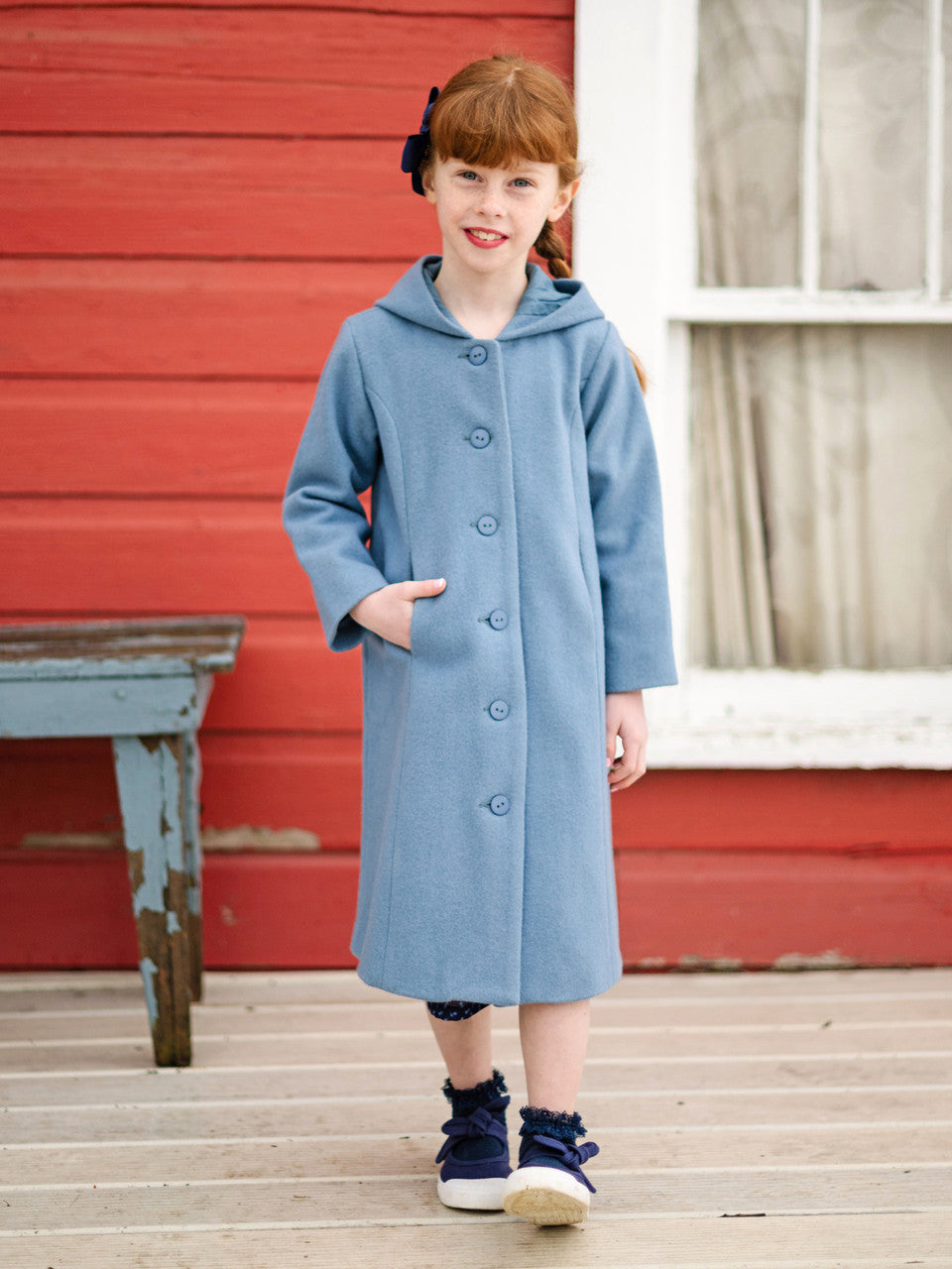 Young girl wearing a blue coat standing on a wooden deck with a red wall in the background.