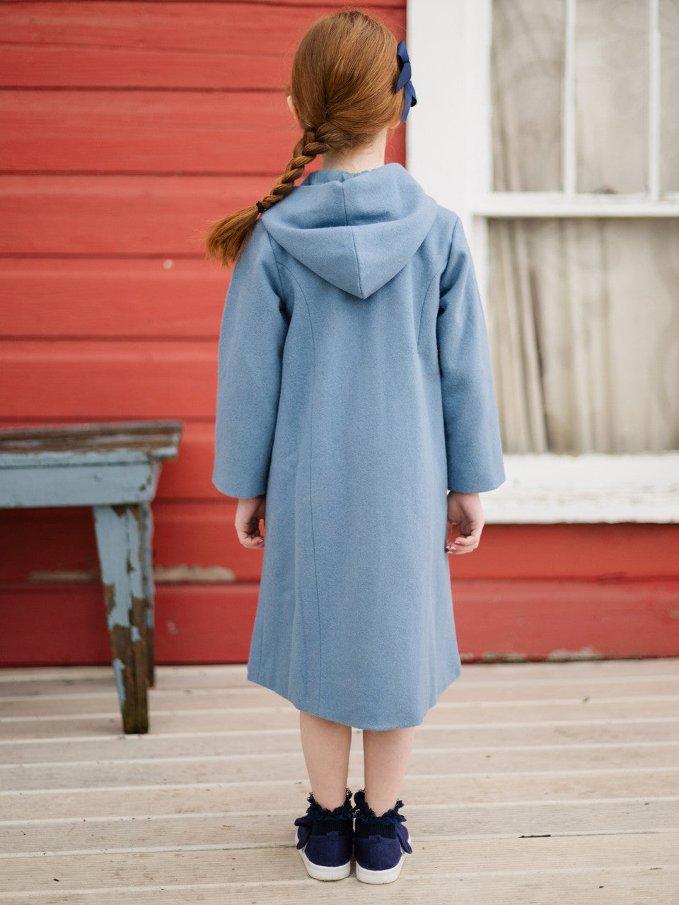 Girl wearing a blue coat standing on a wooden deck with a red wall in the background