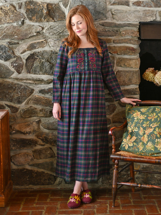 Woman in a plaid dress standing in front of a stone wall with a fireplace.