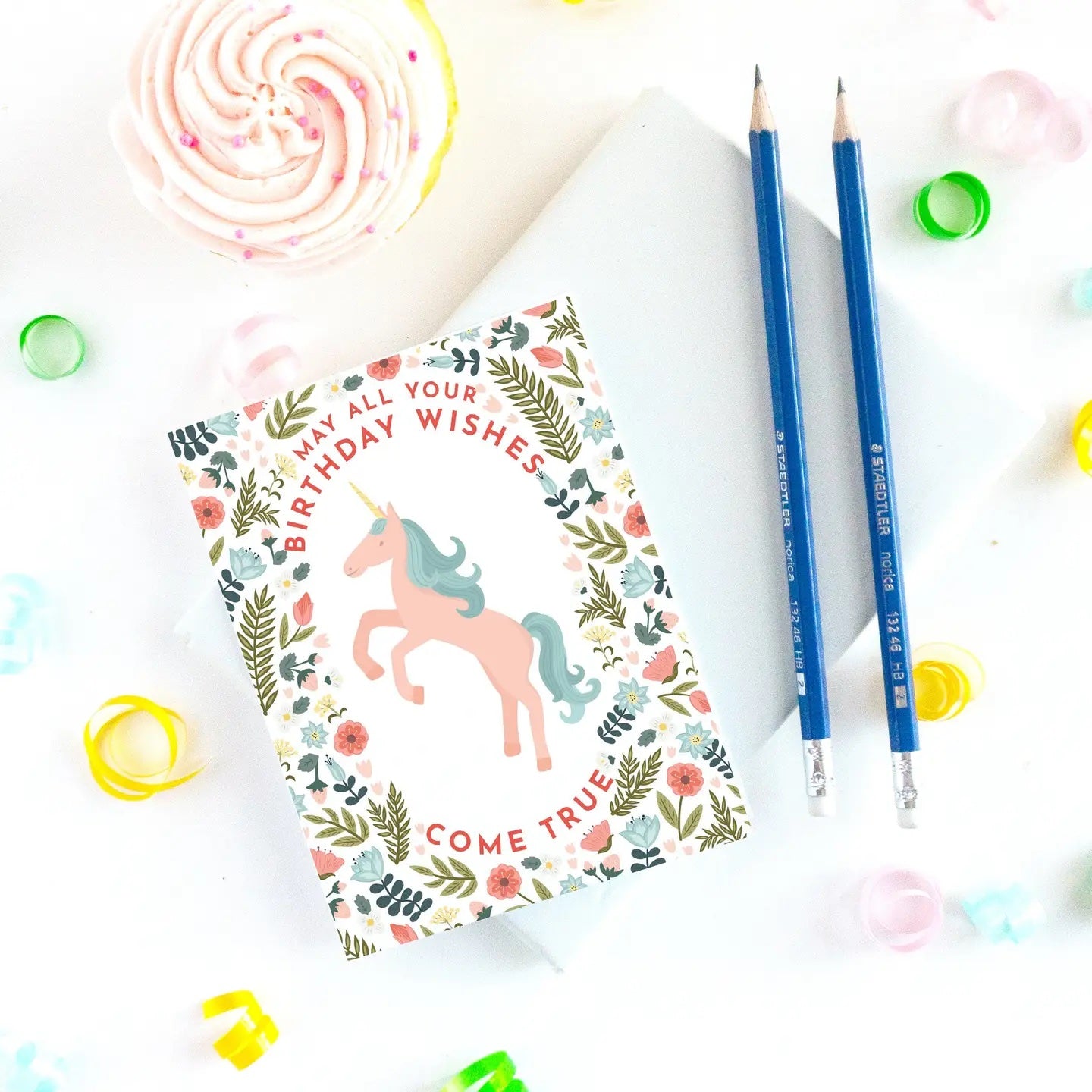 Notebook with unicorn design and birthday wishes text, surrounded by blue pencils and colorful confetti.