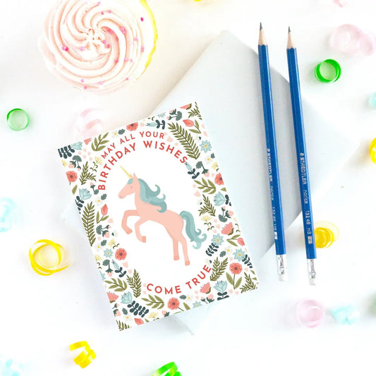 Notebook with unicorn design and birthday wishes text, surrounded by blue pencils and colorful confetti.