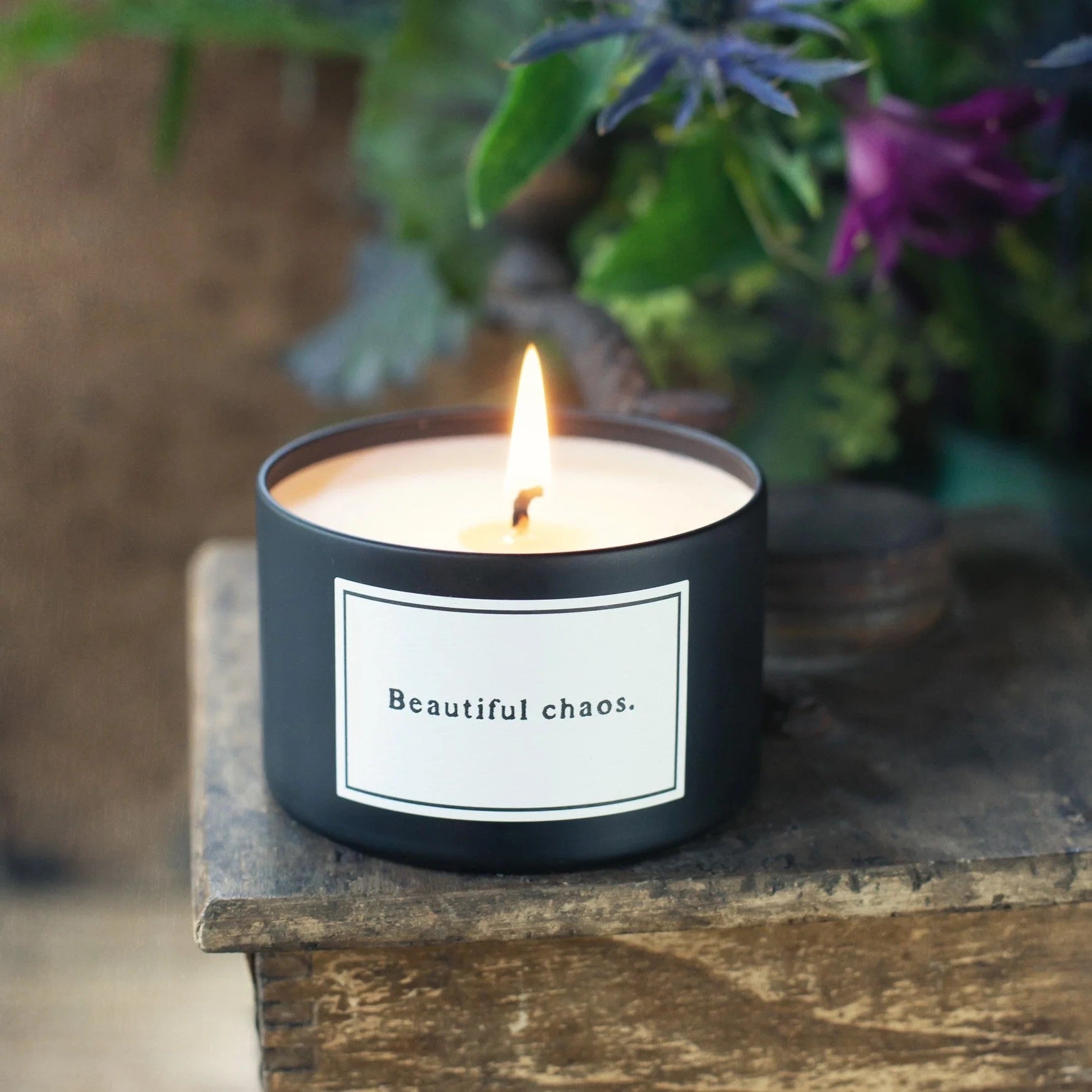 Candle in a black container with a label reading 'Beautiful chaos' on a wooden surface with plants in the background.