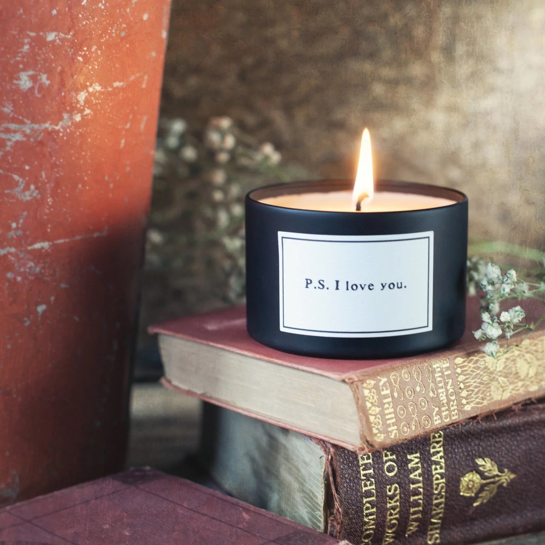 Black candle labeled 'P.S. I love you' on top of vintage books with a blurred background