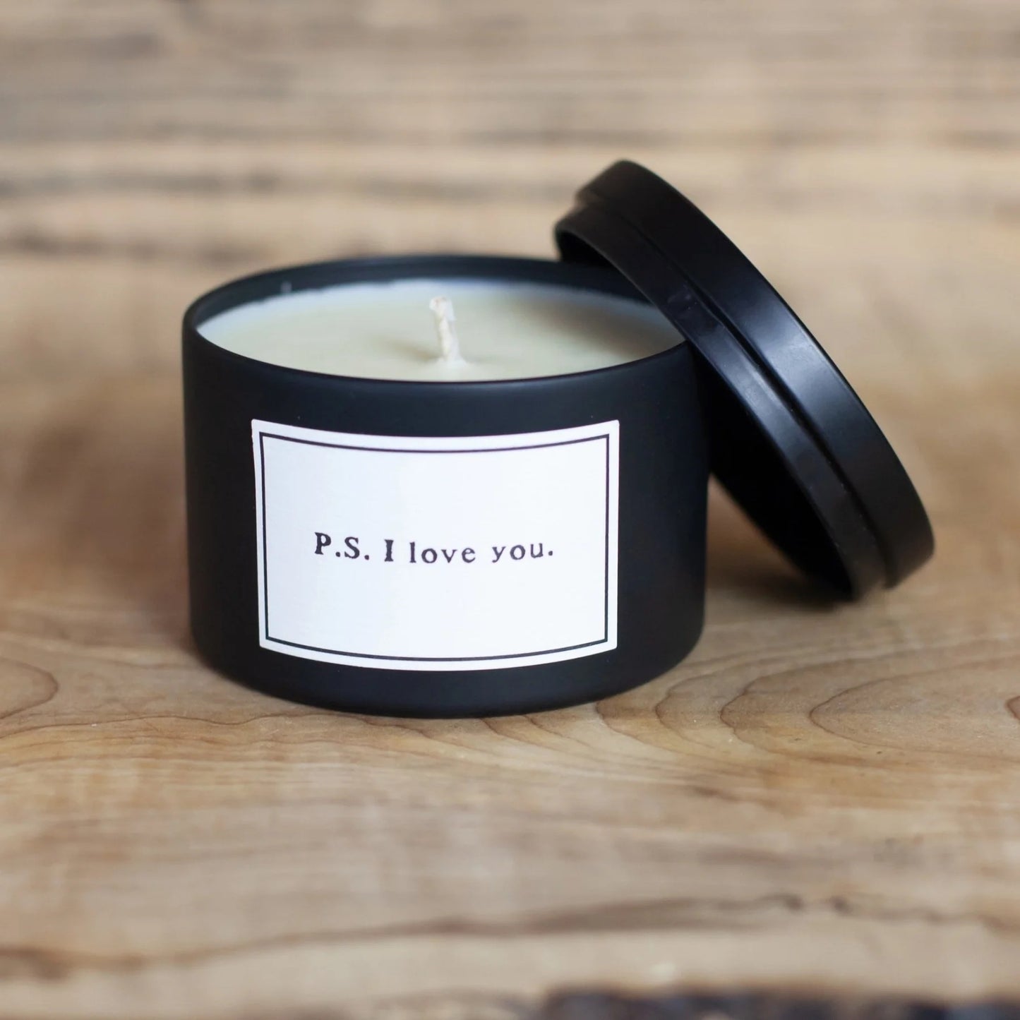 Candle in a black container with a label saying 'P.S. I love you' on a wooden surface