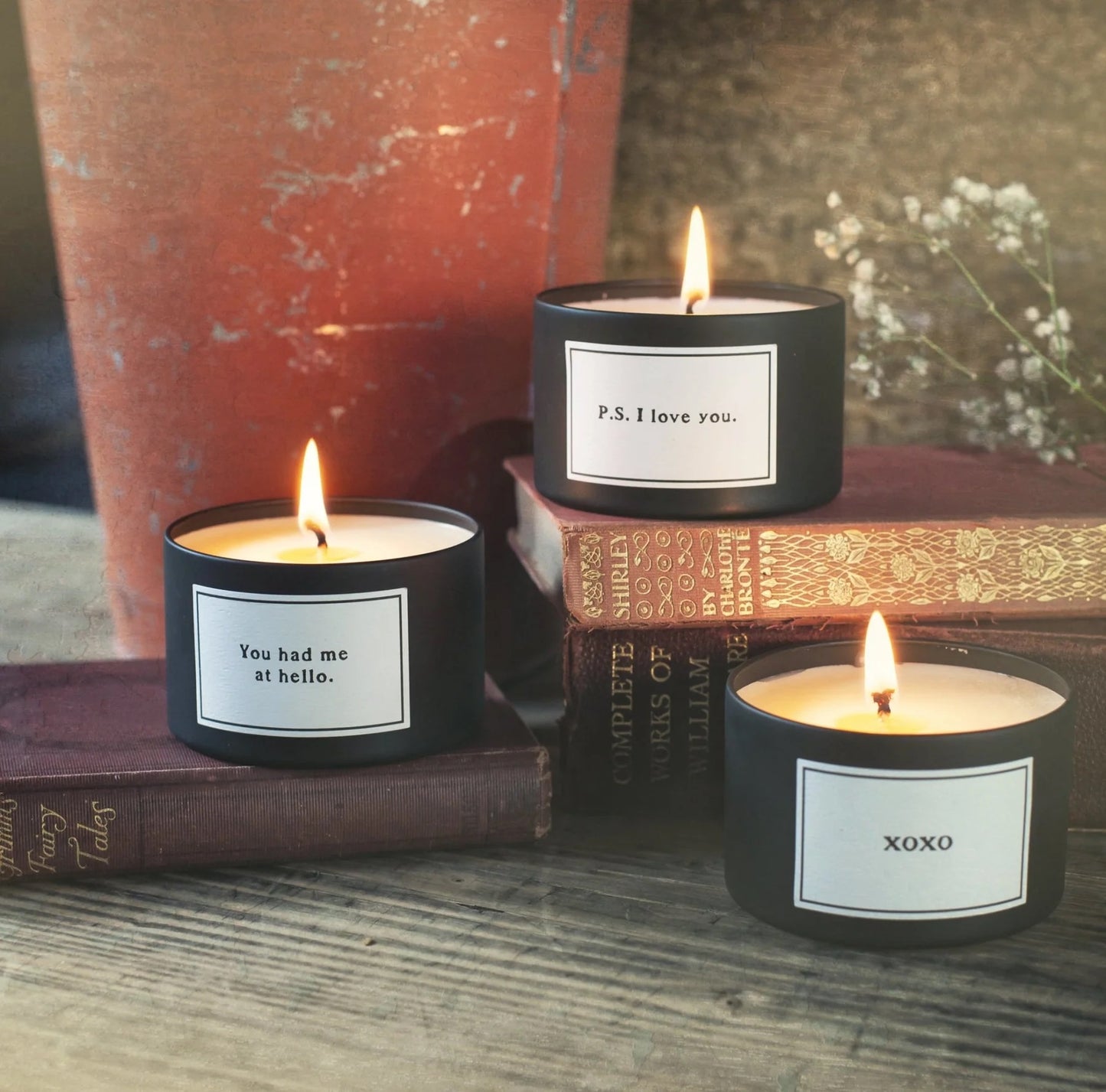 Three lit candles with labels on a wooden surface with books and a plant in the background
