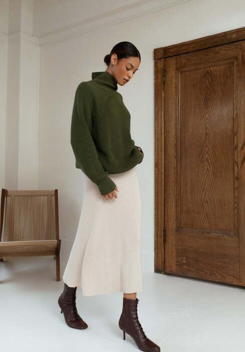 Woman wearing a green sweater and white skirt standing in a room with wooden furniture.